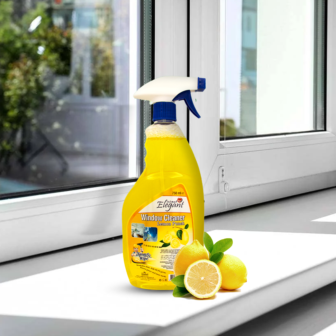 A yellow bottle of Elegant Window Cleaner with a spray nozzle placed on a windowsill, accompanied by fresh lemons in the foreground. A clean, bright window with an outdoor view is visible behind the product.
